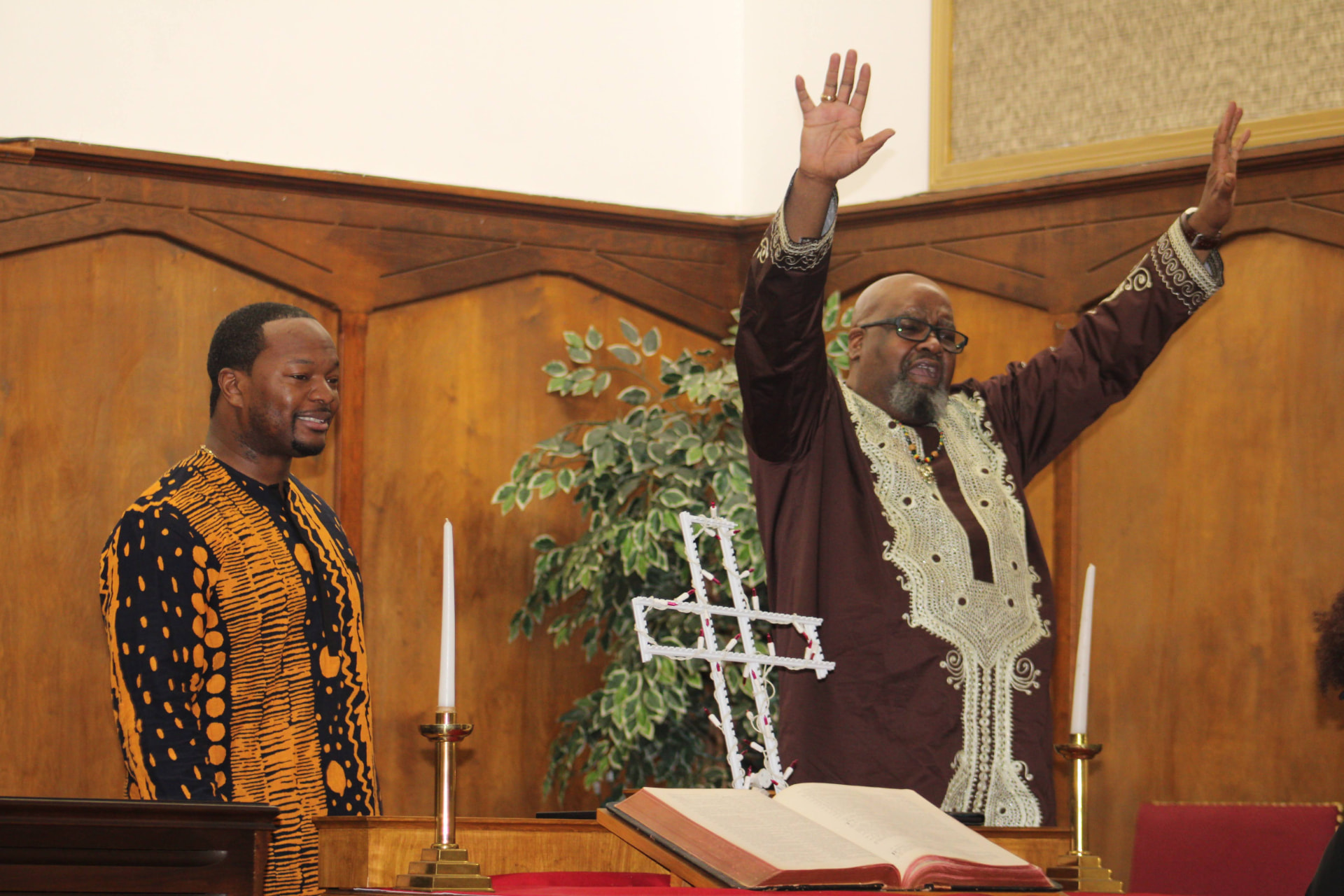Pastor in traditional brown and cream attire raising both hands in praise at the pulpit