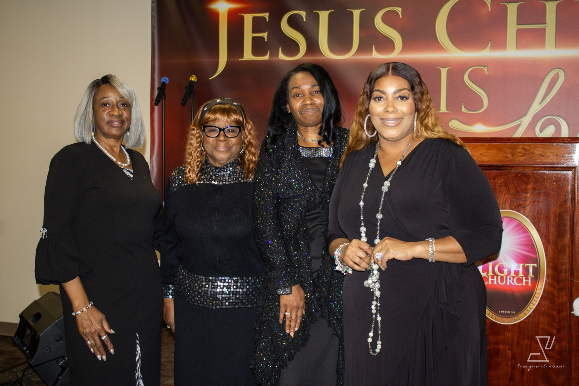 Four women in black posing together beside The Light Church pulpit
