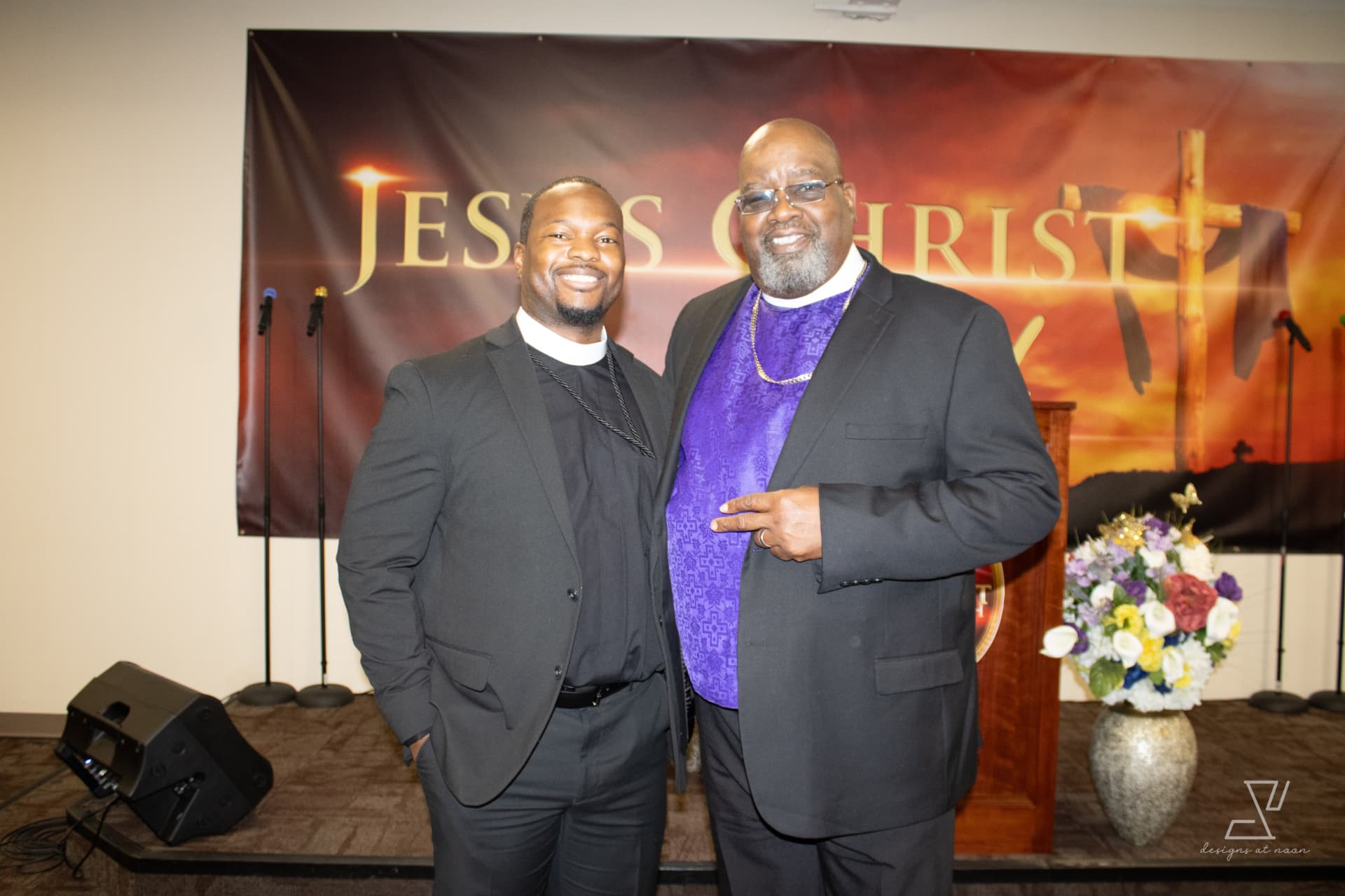 Minister and bishop smiling together in front of the Jesus Christ banner